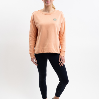 Mulher vestindo sweatshirt cor de pêssego com logo pequeno, leggings pretas e sapatilhas brancas em fundo branco