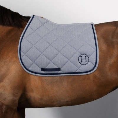 Mantilha para cavalo cinzenta acolchoada com bordos azuis e monograma H, posta numa sela sobre cavalo castanho