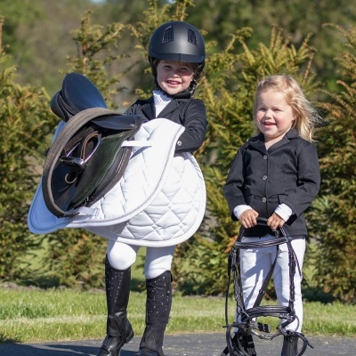Duas crianças vestidas com roupa de equitação segurando equipamento de equitação ao ar livre