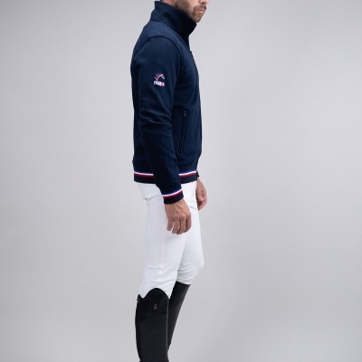 Homem de perfil com casaco azul, calças brancas e botas pretas em fundo cinzento