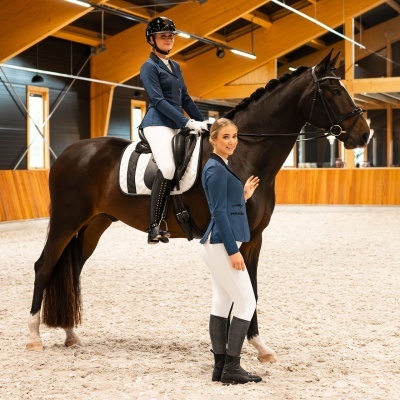 Dois cavaleiros com roupa de equitação formal junto a um cavalo castanho escuro num picadeiro