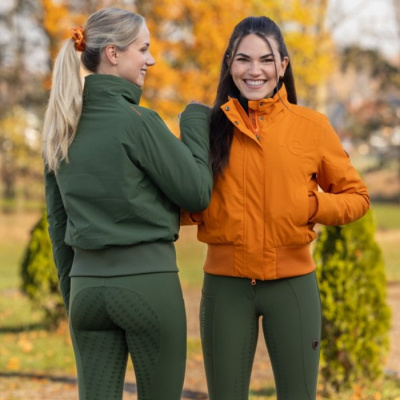 Duas mulheres usam jaquetas verdes e laranja com leggings verdes padrão anatómico em parque outonal.