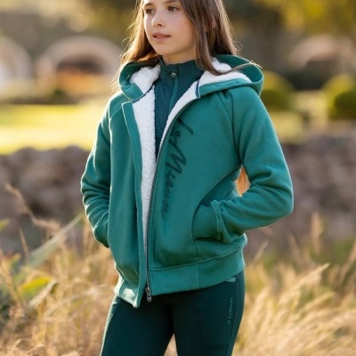 Menina com casaco verde e capuz branco em campo com vegetação seca.