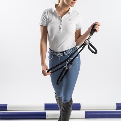 Mulher com roupa de equitação segurando rédeas pretas em fundo branco com barras de salto azul e branco