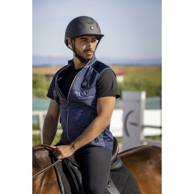 Homem de colete azul e capacete preto sentado a cavalo castanho ao ar livre