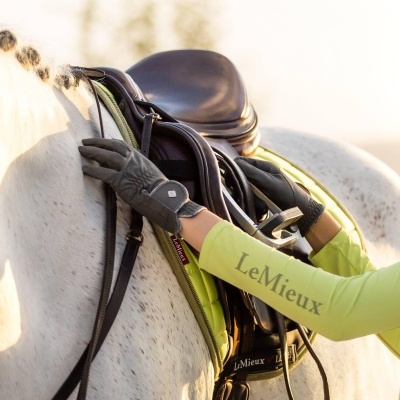 Pessoa a colocar sela castanha num cavalo branco com manta verde-lima e camisa LeMieux