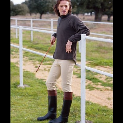 Homem com blusão castanho escuro e calças bege encostado a cerca em área ao ar livre