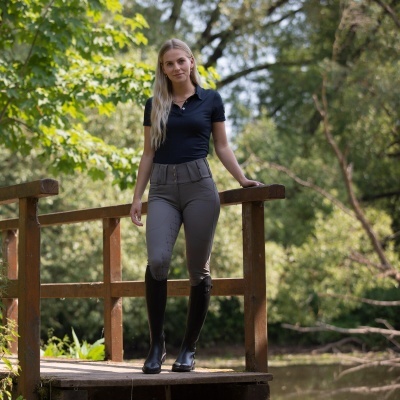 Mulher com camisa preta, calças cinzentas e botas pretas numa ponte de madeira junto a um lago e árvores.