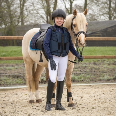 Jovem montador com equipamento de equitação ao lado de cavalo creme com sela e manta azul.
