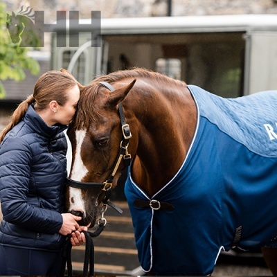 Cavalo castanho com manta azul e pessoa com jaqueta azul escura em ambiente exterior