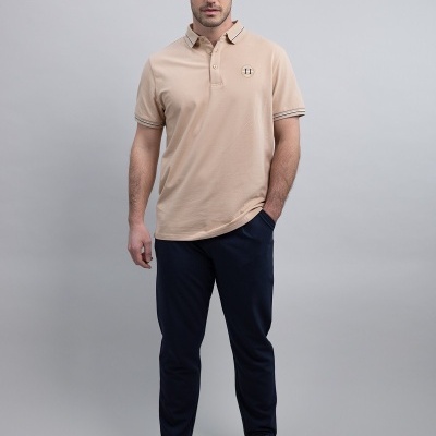 Homem com polo bege, calças azul-escuras e ténis brancos em fundo cinza.