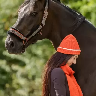 Mulher junto a cavalo com cabresto bege e detalhes em laranja, usando cachecol e gorro laranja.