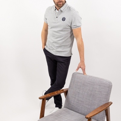 Homem com camisa polo cinza e calças pretas ao lado de cadeira estofada cinza com braços de madeira