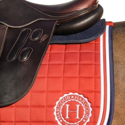 Sela de equitação de couro castanho sobre um pavilhão vermelho acolchoado com bordas azul e branca e logotipo HARCOUR