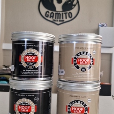 Latas de pomada para cascos Daily Hoof Balm preto e creme empilhadas em superfície branca com logotipo de cavalo ao fundo