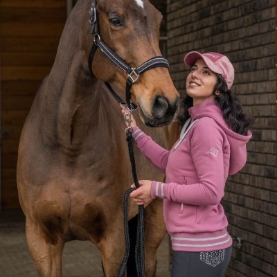 Mulher com casaco de capuz rosa, boné rosa e calças cinzentas ao lado de cavalo castanho com cabeçada preta