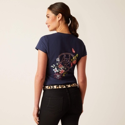 Mulher de costas com t-shirt azul com estampa floral nas costas e calças pretas com cinto de padrão de onça