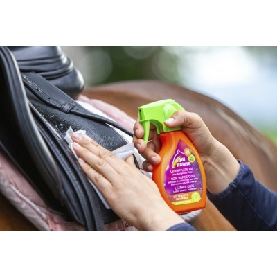 Pessoa a limpar sela de cavalo preta com spray de limpeza, cavalo castanho ao fundo.