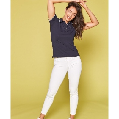 Mulher com polo azul escura, calças brancas e ténis brancos, a posar com os braços levantados contra um fundo amarelo.