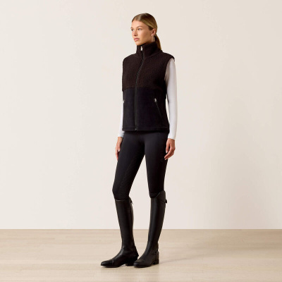 Mulher com colete preto, t-shirt branca, leggings pretas e botas pretas em ambiente interior claro