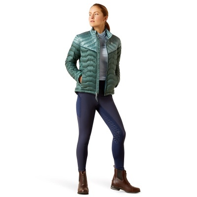 Mulher com casaco verde acolchoado, leggings azuis e botas castanhas
