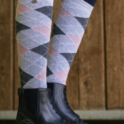 Meias compridas com padrão de losangos cinzentos e rosa, botas pretas de pele, fundo de madeira