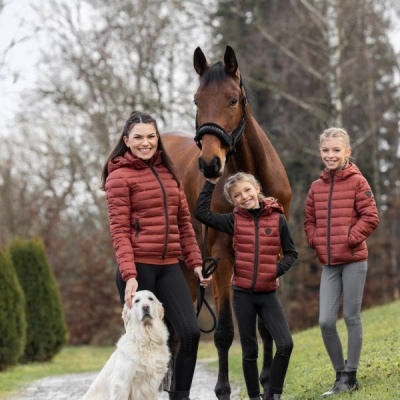 Mulher e duas crianças com casacos e coletes acolchoados cor ferrugem junto a um cavalo castanho e um cão branco ao ar livre