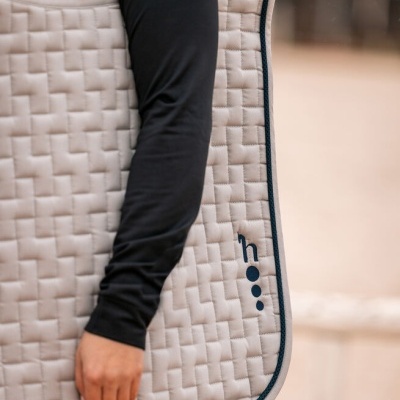 Tapete de sela bege matelassê com logótipo 'h...' azul escuro segurado por pessoa com camisa preta.