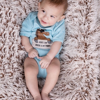 Bebé em body azul claro com estampa de cavalo castanho, deitado em tapete felpudo bege claro