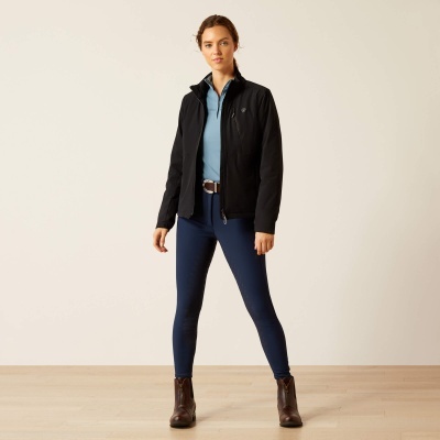 Mulher com casaco preto, camisa azul clara, calças azuis e botas castanhas em ambiente interno