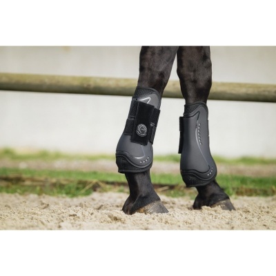 Botas de proteção pretas em patas traseiras de cavalo castanho escuro em piso de areia.