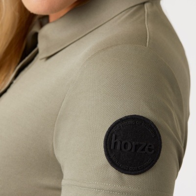 Polo verde-oliva com emblema preto na manga e mulher loira ao fundo branco