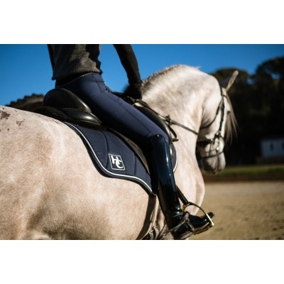 Cavalo branco com sela preta e alforge azul escuro com borda branca e logótipo branco, cavaleiro em calças de equitação e botas pretas