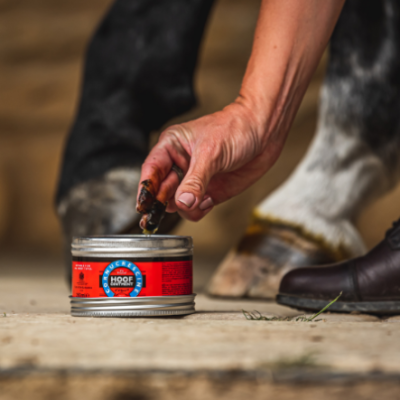 Lata de produto para cascos de cavalo com mão a retirar o produto e patas de cavalo ao fundo