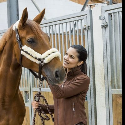 Mulher com blusão castanho a segurar rédea de cavalo castanho com arreio acolchoado branco em estábulo