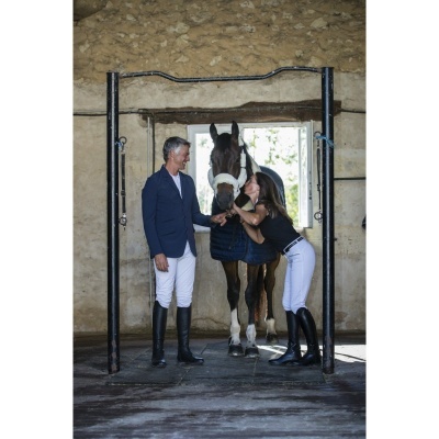 Cavalo castanho com peitoral e manta azul entre duas pessoas com roupa de equitação em espaço interior