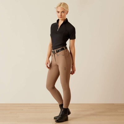 Mulher com camisa preta de manga curta, calças justas castanhas, cinto preto e botins pretos, em interior com chão madeira e parede bege