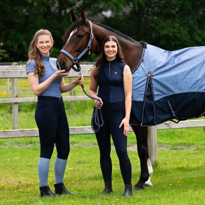 Duas mulheres com roupa de equitação e cavalo com manta azul clara e cabresto azul na relva