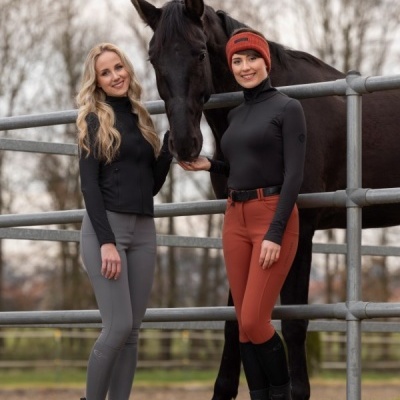 Duas mulheres com roupa de equitação e um cavalo negro ao ar livre junto a uma cerca metálica
