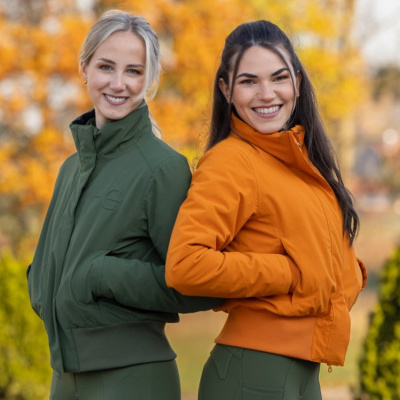 Duas mulheres com casacos verde escuro e laranja em ambiente exterior com árvores amarelas
