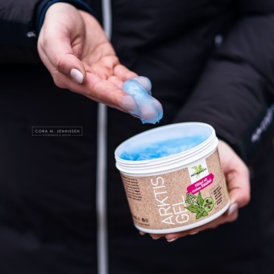 Frasco de gel azul Arktis Gel com tampa branca e rótulo castanho, mãos segurando e espalhando gel.