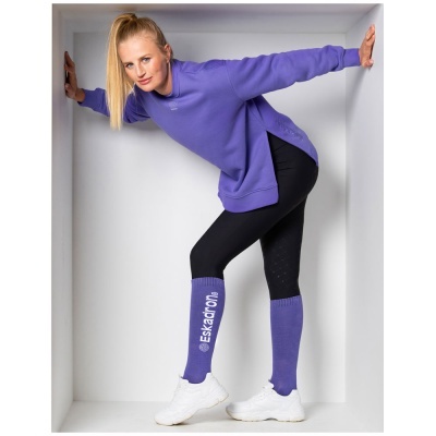 Mulher com sweatshirt roxa, leggings pretas e polainas roxas com texto branco numa caixa branca