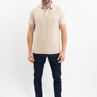 Homem com polo bege, calças jeans azul-escuro e ténis brancos em fundo branco