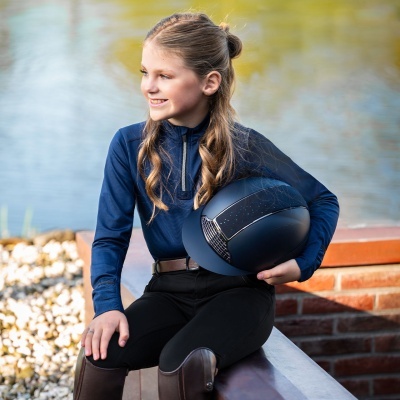 Menina com roupa de equitação azul e preta segurando capacete preto sentada junto a muro de tijolos perto de lago.