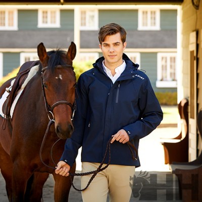 Homem jovem com casaco azul escuro e cavalo castanho com sela branca junto a construção rústica