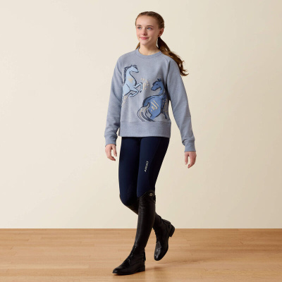 Mulher jovem a usar sweatshirt cinzenta com estampado de cavalos, leggings azuis escuras ARIAT e botas pretas de equitação