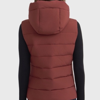 Colete acolchoado vermelho com capuz, usado com camisola preta, jeans e boné, visto de costas