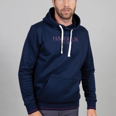 Homem com hoodie azul escuro com cordões brancos e texto rosa, calças cinza, fundo cinza