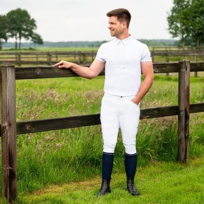 Homem com roupa branca de equitação e botas pretas encostado numa vedação de madeira num campo verde