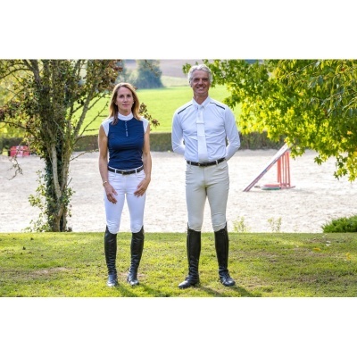 Duas pessoas vestidas com roupa de equitação estão ao ar livre em pé sobre relva com fundo de areia e árvores.